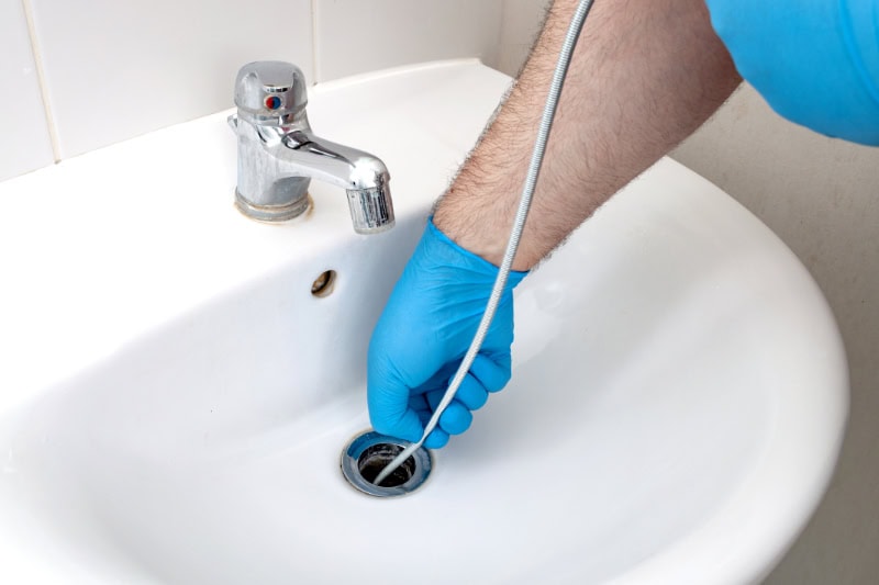 4 Reasons to Clean Your Drains Before the Holidays. Plumbing issues, occupation in sanitation and handyman contractor concept with plumber repairing drain with plumbers snake (steel spiral that twists through pipes to collect dirt) in residential sink.